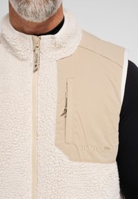 Beige fleece vest med en blød, struktureret front, glatte stofaccenter, en lynlåslomme og broderet logo på siden.
