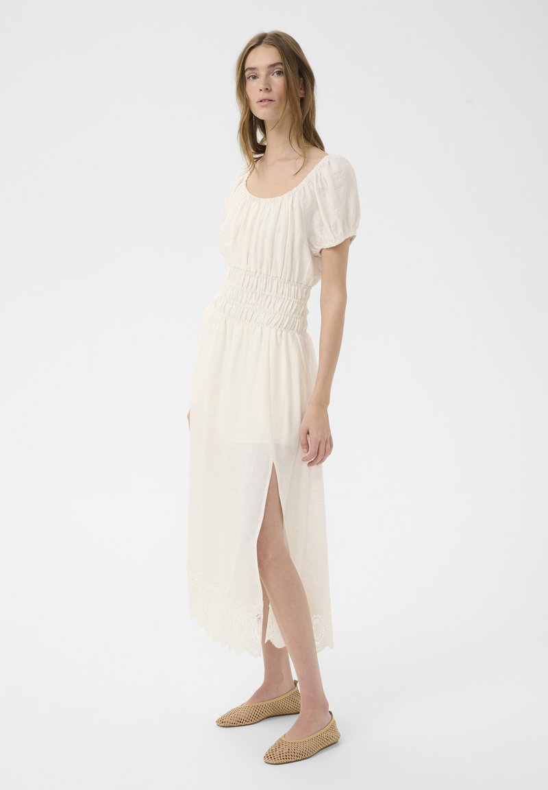 Jeune femme portant une robe midi blanche à manches bouffantes et fente latérale, associée à des ballerines tissées beiges, debout sur un fond blanc.