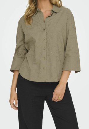 Kobieta ubrana w luźną oliwkową bluzkę na guziki z kwiatowym haftem i rękawami do łokcia, w połączeniu z czarnymi spodniami i złotymi pierścionkami.