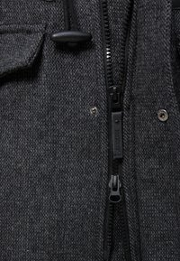 Sezione di giacca grigia con trama, caratterizzata da una cerniera centrale nera, bottoni a pressione e un cordoncino regolabile con fermo. Vista ravvicinata che evidenzia la tessitura del tessuto.