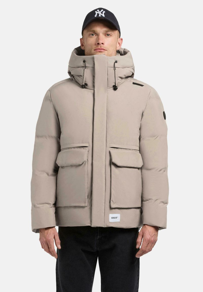 Light beige puffer jacket with a high collar, drawstring hood, and front flap pockets. Features black accents and a brand label at the hem.