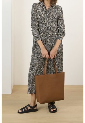 Frau in einem langärmeligen schwarz-weiß gemusterten Kleid, die eine große braune Ledertasche hält und schwarze gewebte Sandalen auf einem Holzboden trägt.