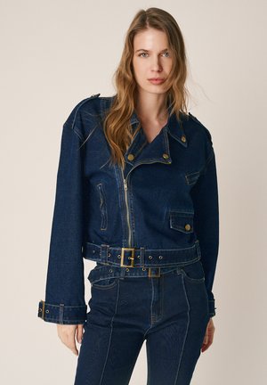 Frau mit langen hellbraunen Haaren, die eine dunkelblaue Jeansjacke mit Gürtel und dazu passende hoch taillierte Jeans vor einem schlichten Hintergrund trägt.