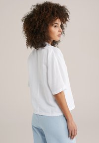 Top bianco in lino a maniche corte con una vestibilità morbida, caratterizzato da un collo rotondo e un dettaglio di cucitura sul retro. Abbinato a pantaloni a righe blu chiaro.