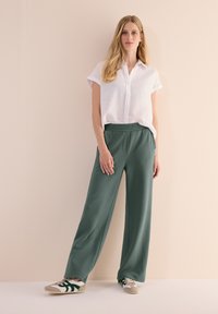Losse groene broek met een gestructureerd patroon, gecombineerd met een witte kortebloes met knopen en beige sneakers met groene accenten.
