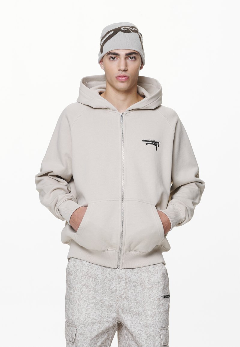 Beige Zip-Hoodie mit Kängurutaschen, ausgestattet mit einem schwarzen grafischen Detail auf der Brust und einer weichen Textur. Kombiniert mit gemusterten hellen Hosen.