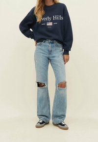 Persona che indossa una felpa blu navy con la scritta "Beverly Hills", jeans strappati di colore blu chiaro con buchi alle ginocchia e sneakers nere e grigie, su uno sfondo chiaro.
