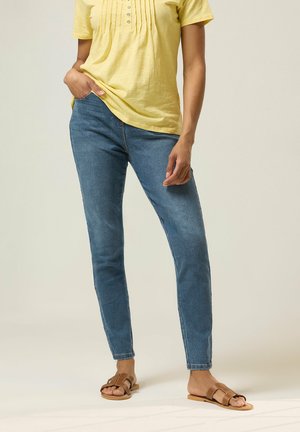 Frau trägt gelbes Kurzarmshirt, blaue eng anliegende Jeans und braune Pantoletten, steht mit einer Hand in der Tasche vor einem schlichten Hintergrund.