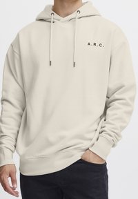 Sweat à capuche couleur crème avec une poche frontale, une capuche à cordon réglable, et un petit logo noir "A.R.C." sur la poitrine gauche. Texture douce.