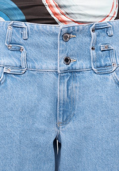 Ανοιχτό μπλε τζιν ψηλόμεσο με φαρδιές θηλιές για τη ζώνη, δύο μαύρα κουμπιά και μεταλλικά καρφιά για κούμπωμα στο μπροστινό μέρος.