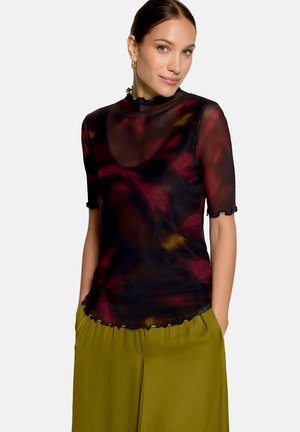 Femme avec les cheveux attachés porte des boucles d'oreilles créoles dorées, un haut noir transparent avec des motifs rouges et jaunes, et un pantalon ample vert olive avec les mains dans les poches.