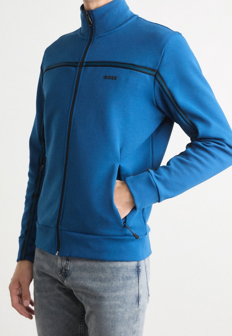 Blue zip-up jacket with a high collar, black accents, side pockets, and a fitted design. Material appears soft and stretchy.