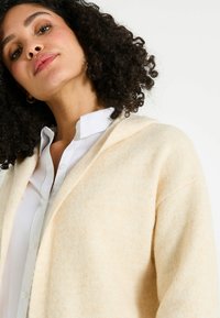 Cardigan beige clair avec une texture douce, design en V et ouvert à l'avant. Porté sur une chemise blanche à boutons, avec des épaules tombantes.