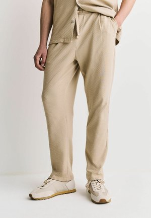 Mann trägt beige strukturierte Hose, passenden beigen Oberteil und hellbeige Sneakers, steht vor einem schlichten hellen Hintergrund.