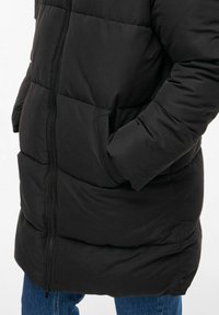 Cilvēks valkā melnu pārklātu ziemas jaku ar priekšējo rāvējslēdzēju un rokām kabatās, komplektā ar ziliem džinsiem.