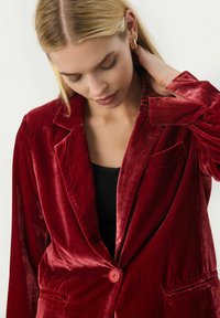 Blazer in velluto rosso con rever a punta, chiusura a un bottone e dettagli delle tasche. Texture liscia, vestibilità rilassata. Indossato sopra una maglietta nera.
