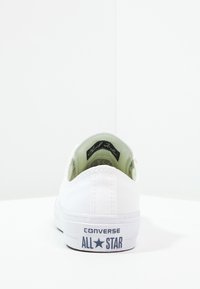 Vit canvas lågskor med gummisula, med en svart "Chuck Taylor"-etikett inuti och "CONVERSE ALL STAR"-logotyp på hälen.