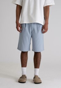 Light blue shorts with a loose fit and soft texture, featuring two front pleats and side pockets, paired with a white oversized T-shirt and beige clogs.