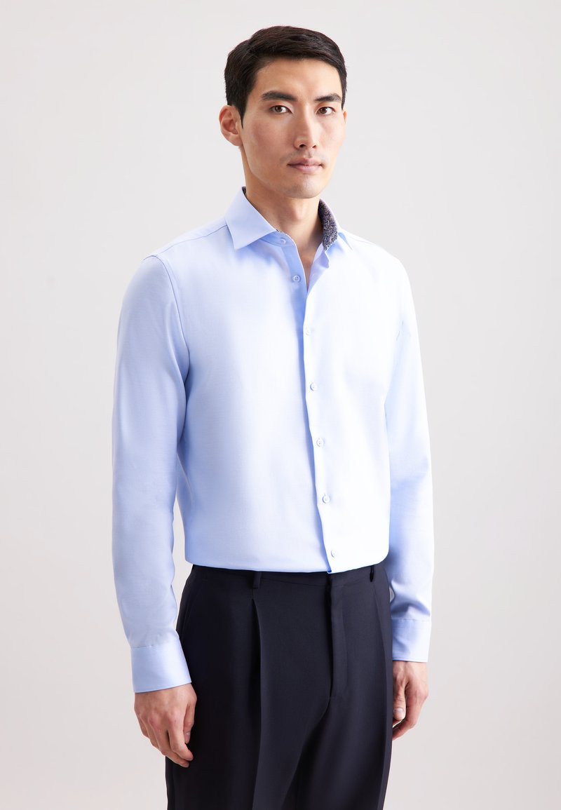 Camicia azzurra chiara con una texture morbida, colletto a punta e colletto interno a fantasia in contrasto, abbinata a pantaloni scuri plissettati.