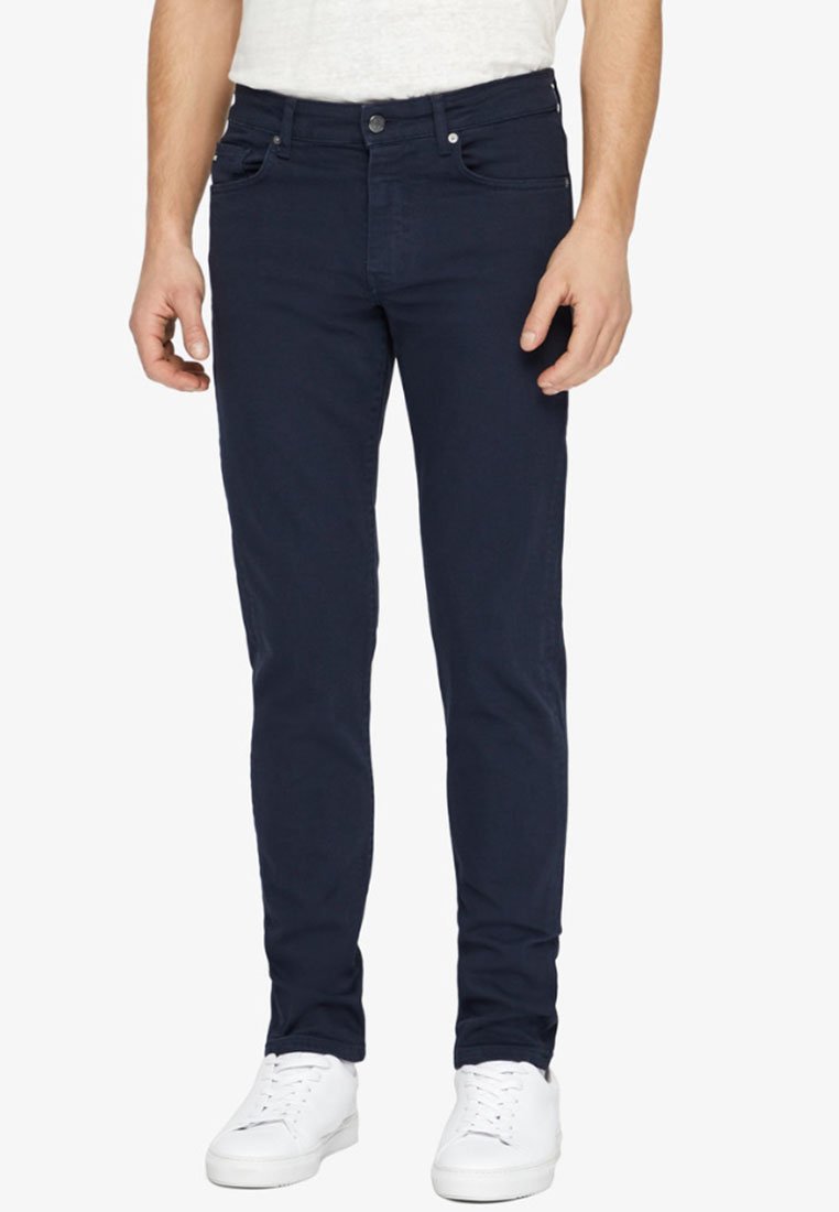 Jeans slim-fit de culoare albastru marin, cu o textură netedă, dotate cu un sistem de închidere cu buton și fermoar, buzunare frontale și un design minimalist, purtate cu adidași albi.
