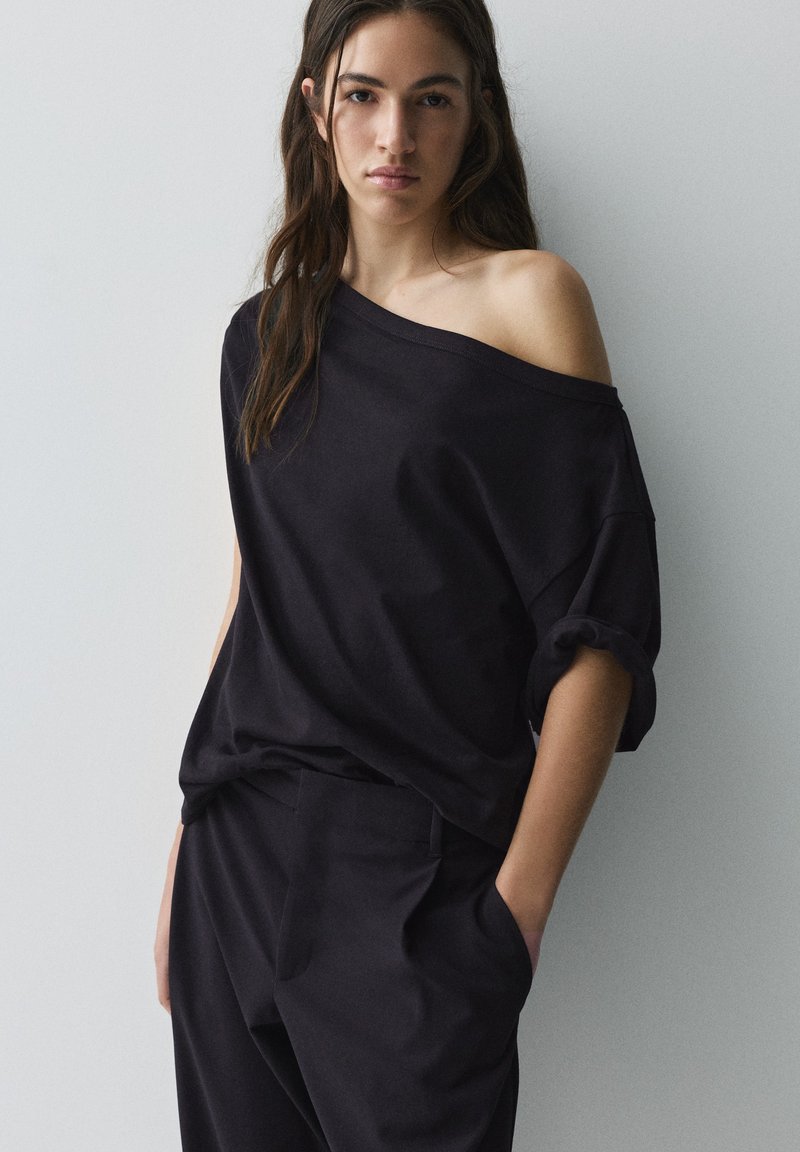 Femme aux longs cheveux foncés portant un haut noir décolleté sur les épaules et un pantalon noir, debout avec une main dans la poche sur un fond uni.