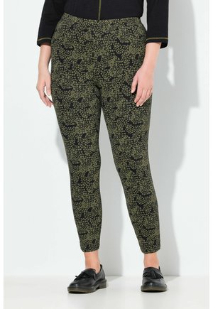Donna che indossa pantaloni aderenti neri e oliva a stampa, mocassini neri con frange e un top lungo nero, in piedi contro un muro bianco.
