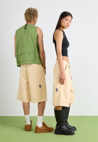 Due modelli stanno schiena contro schiena. Uno indossa un gilet senza maniche verde, pantaloni corta beige e sneakers marroni. L'altro indossa un crop top nero, pantaloni corta beige e stivali neri fino al ginocchio.