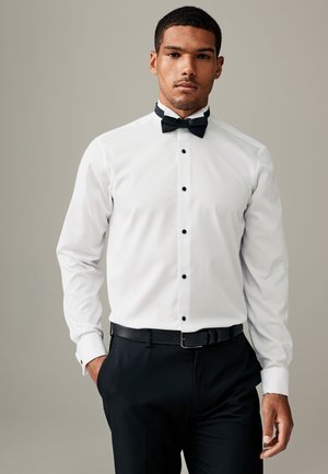 Uomo che indossa una camicia da smoking bianca con bottoni neri e papillon, cintura nera e pantaloni eleganti neri, in piedi su uno sfondo semplice.