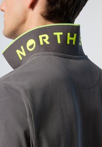 Giacca grigia con collo alto, rifinita con un bordo verde brillante e la scritta "NORTH", che mette in evidenza un tessuto testurizzato e un design strutturato.