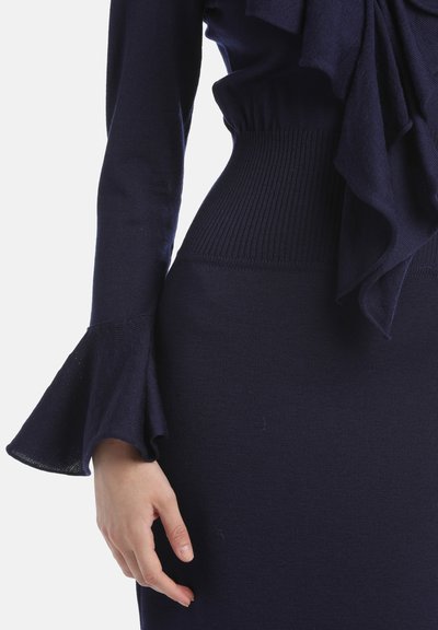 Temno modra pletena obleka s fitted korpusom, razširjenimi rokavi in ribastim pasom. Gladka tekstura z plastnim volanom na dékolteju.