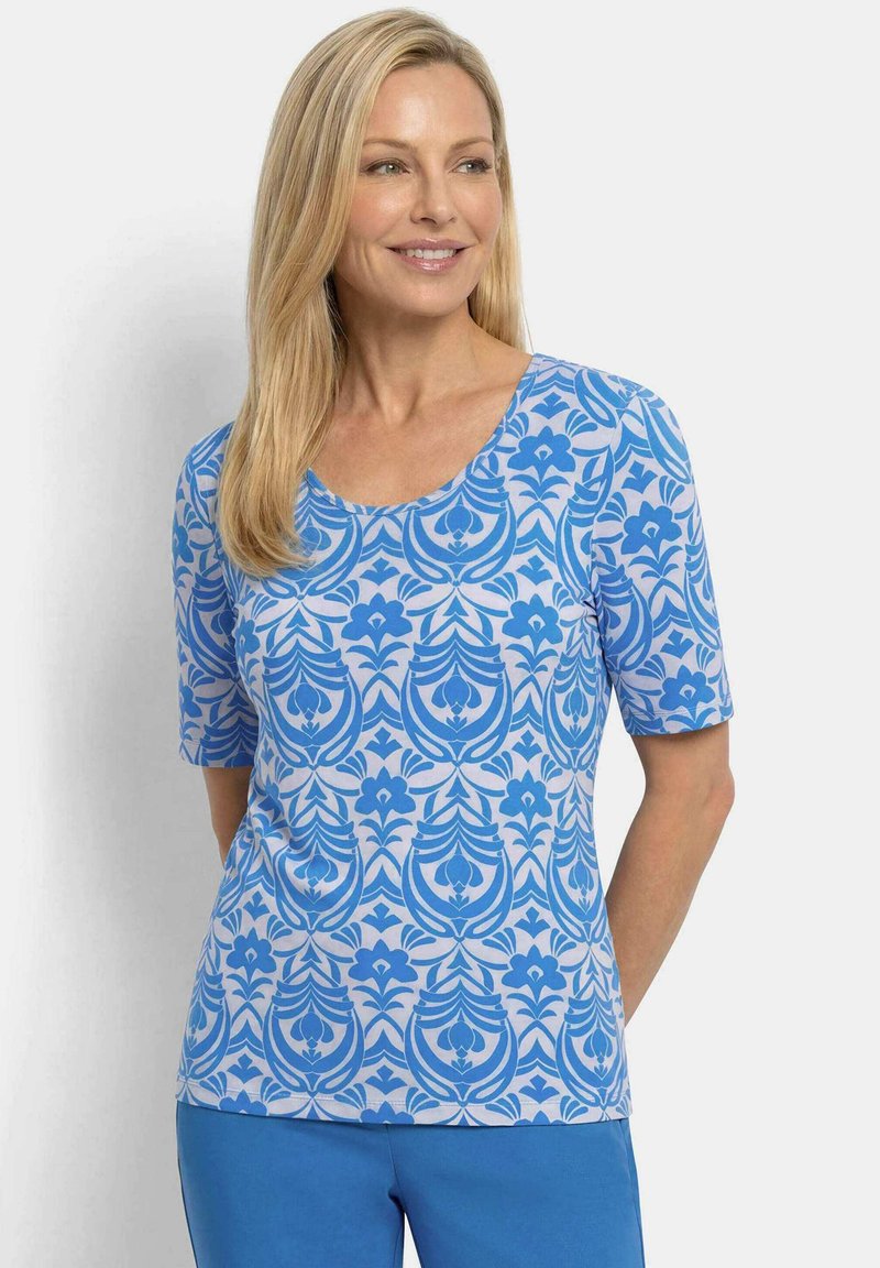 Femme aux cheveux blonds portant un haut à manches courtes à motif floral bleu et blanc et un pantalon bleu uni, regardant sur le côté avec un sourire.