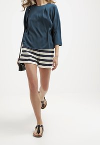 Ciemnozielony, luźny top z rękawami 3/4, w zestawieniu z szortami w paski. Modelka nosi czarne klapki i trzyma czarną torbę na ramię.