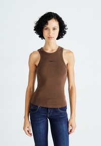 Vrouw met krullend haar, gekleed in een bruine mouwloze ribgebreide top en donkerblauwe jeans, staand tegen een effen witte achtergrond.