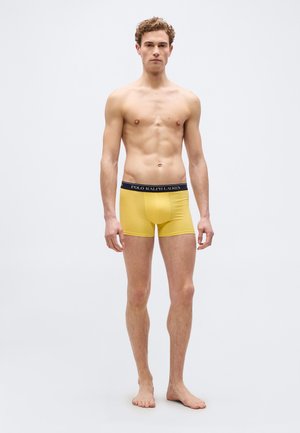 Tânăr stând desculț, purtând chiloți boxer galbeni cu talie neagră inscripționată cu „Polo Ralph Lauren”, pe un fundal alb simplu.