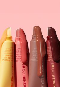 Cinco tubos de tratamiento labial en amarillo, rosa, beige, marrón y marrón oscuro, con acabados brillantes y etiquetas de texto en colores contrastantes.