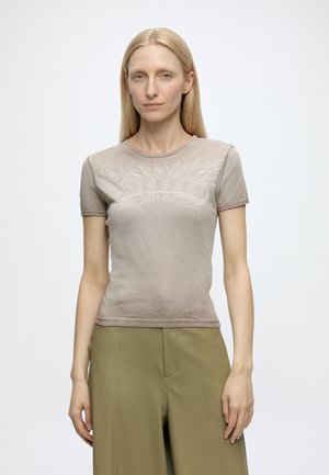 Femme aux cheveux blonds et lisses portant un T-shirt Diesel taupe ajusté et un pantalon vert olive, debout devant un fond blanc uni.