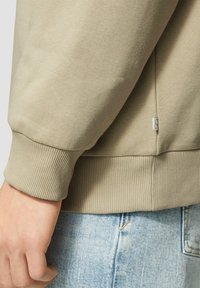 Felpa beige con polsini e orlo a coste, texture morbida e una piccola etichetta con logo vicino alla cucitura laterale. Indossata con denim azzurro chiaro.