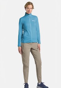 Veste polaire bleu clair à fermeture éclair avec manches longues, dotée d'un col montant et de poches latérales. Associée à un pantalon beige et des chaussures de sport bleues.