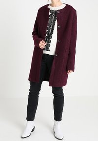 Abrigo de lana burdeos con corte recto, cuello redondo y cinco grandes botones negros; combinado con un suéter blanco y pantalones negros.