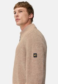 Beige Strickpullover mit hohem Kragen, strukturiertem Stoff und einem schwarzen Patch mit gelb-weißem Logo auf dem linken Ärmel.