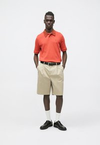 Oranžové polo tričko, khaki šortky, černý pásek, černé boty a bílé ponožky. Model stojí s rukama v kapsách na jednobarevném pozadí.