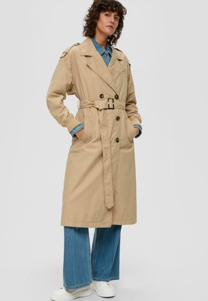 Manteau beige à fermeture bouton, ceinture et poches latérales. Fabriqué en tissu léger. Associé à une chemise en denim et à un jean à jambes larges.
