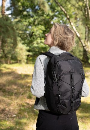 Mochila negra que presenta un tejido texturizado, forma curva, correas ajustables y detalles de cordón elástico. Llevada por una persona en un entorno boscoso.