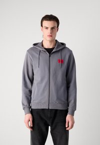 Siva zip-up dukserica s kapuljačom, s crvenim logotipom na lijevoj strani prsnog koša, izrađena od glatkog, teksturiranog materijala. Uparena s crnim trapericama.