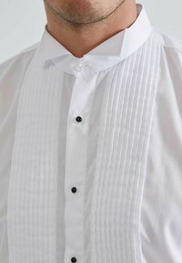 Chemise blanche à col haut avec un devant plissé, ornée de boutons noirs. Texture lisse, coupe ajustée et sans motifs visibles.