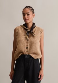 Tan knitted sleeveless sweater with a V-neck, textured fabric, and two front buttons, paired with a black scarf and high-waisted black pants.