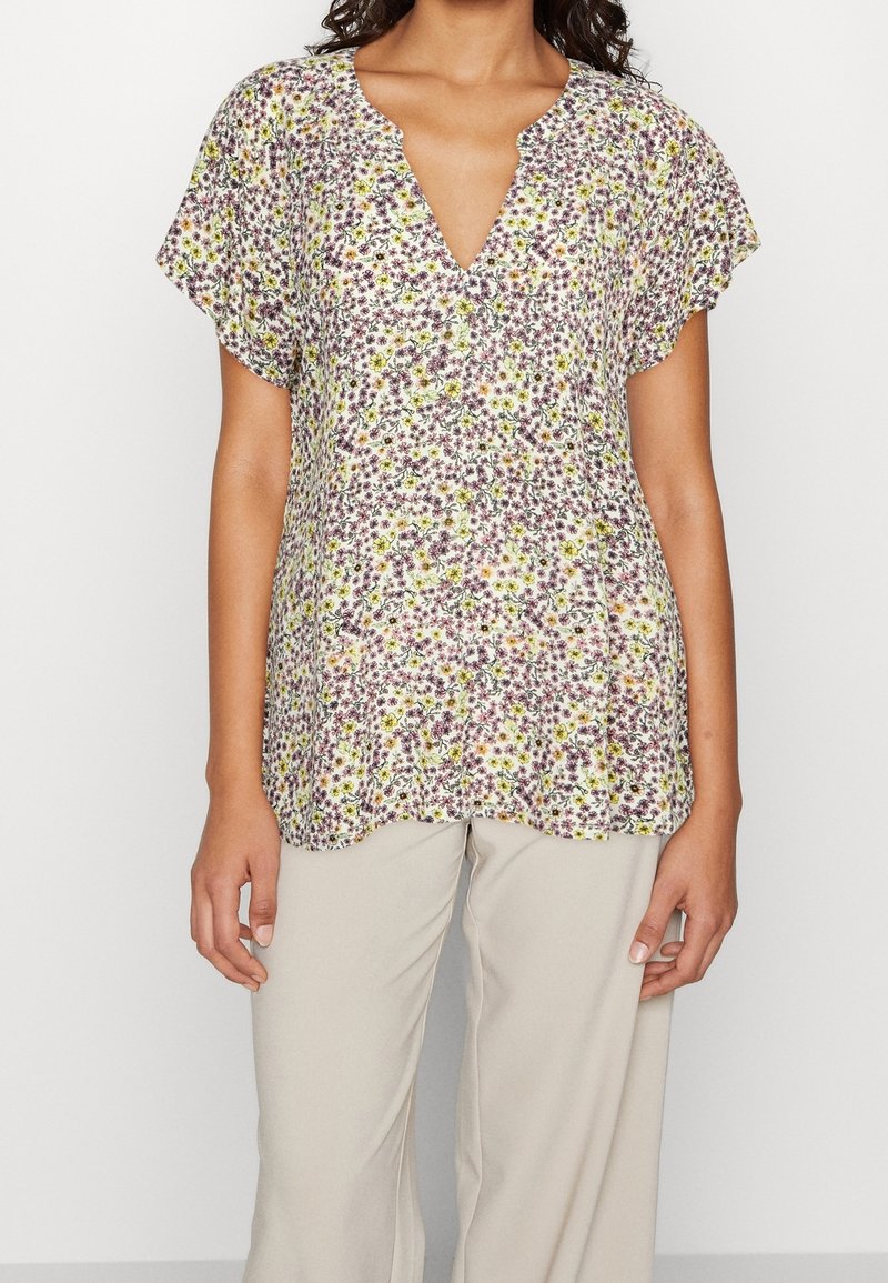 Mujer vestida con una blusa floral de manga corta con flores púrpuras y amarillas, combinada con pantalones beige claros, frente a un fondo liso.