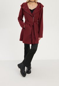 Abrigo burdeos con capucha, cinturón y cierre de cremallera. Mangas largas y bolsillos laterales. Llevado con pantalones ajustados negros y botas negras gruesas.