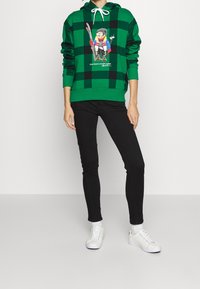 Zelená kostičkovaná mikina s předním vzorem kresleného medvěda v lyžařském vybavení. Kombinována s černými slim-fit džíny a bílými teniskami.