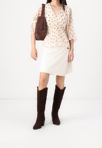 Top floral crema con mangas abullonadas, falda blanca texturizada y botas de ante marrón hasta la rodilla. Un bolso de hombro marrón añade contraste.